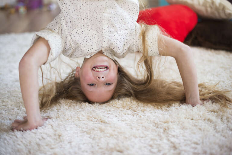 A cheerful young girl with long blonde hair doing a headstand on a soft, plush rug in a cozy living room.