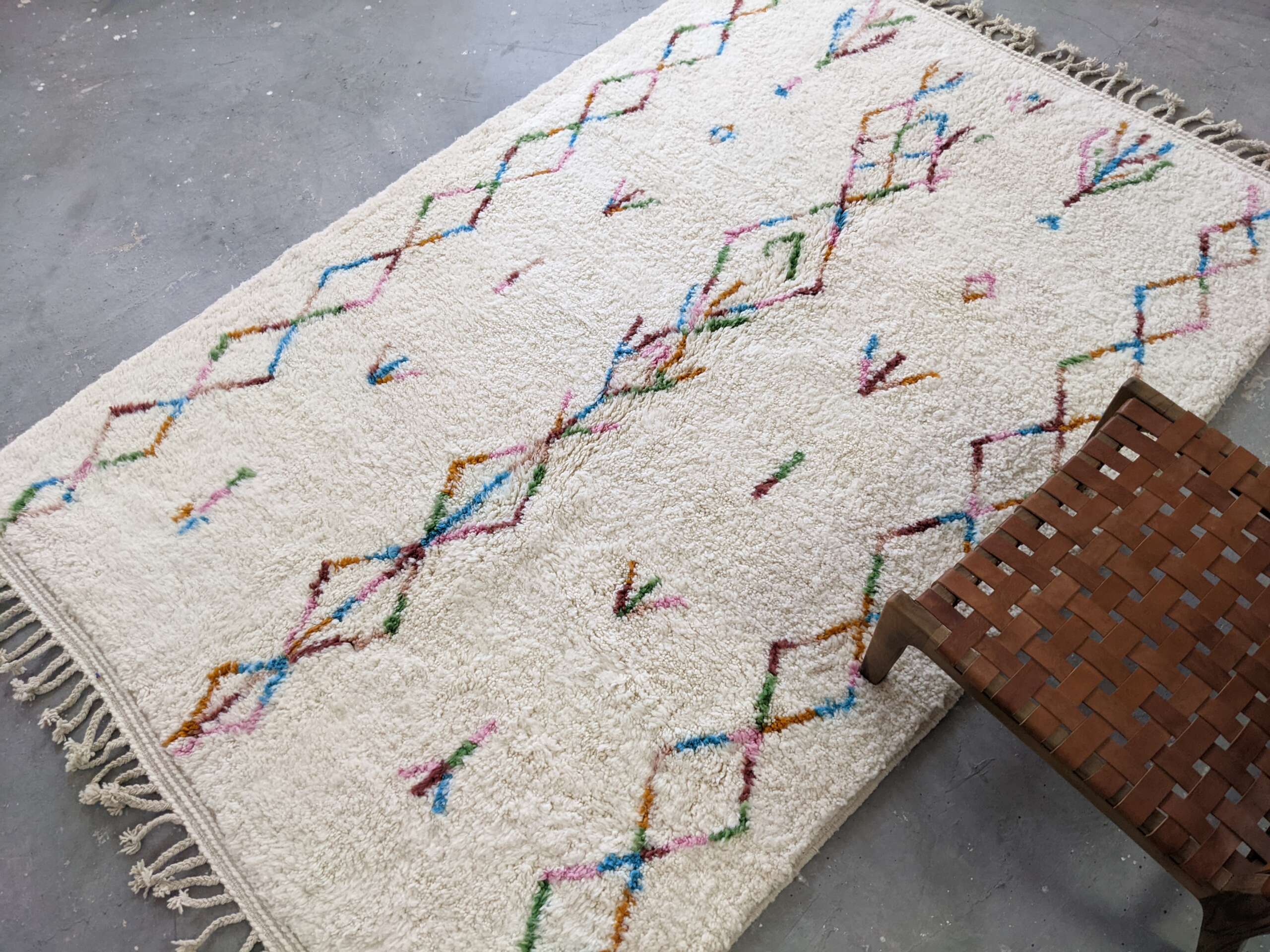 A handmade Moroccan Berber rug with colorful diamond patterns on a soft cream background, placed on a concrete floor near a woven leather stool.