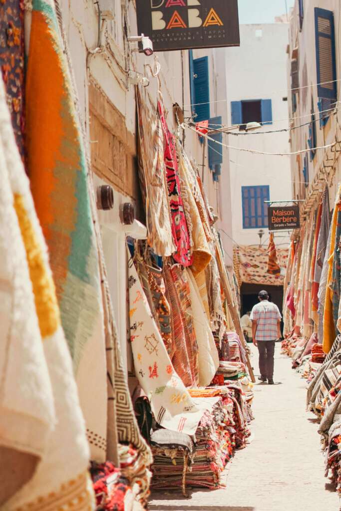A narrow Moroccan street lined with colorful handmade Berber rugs displayed on walls and stacked on the ground. A man walks through the alley, surrounded by traditional textiles and carpets.