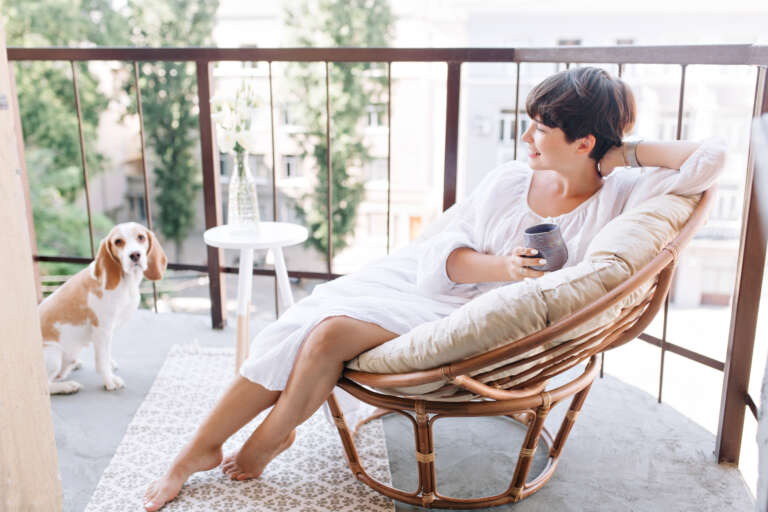 A woman in a white dress sits barefoot in a cozy rattan chair on a balcony, sipping tea and enjoying the outdoor view. A beagle dog stands nearby, looking towards the camera. A patterned rug, small side table with a vase of flowers, and a metal railing complete the setting.